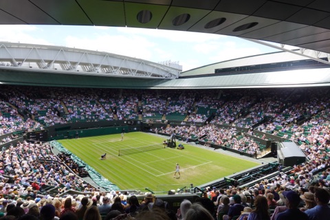 A grass tennis court seen from high in the stand during a match, the retractable roof open above it and every seat in the ground filled