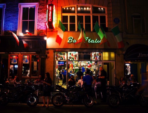 Bar Italia's front at night: green neon script over the window, Italian flags along the fascia, the lit counter inside crowded with people, and motorbikes parked along the kerb.