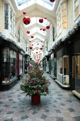 The arcade at Christmas: a tree hung with red baubles standing on the patterned marble floor, rows of large red baubles suspended the length of the glass roof, frosted garlands along the black shopfronts and a lit window display either side.