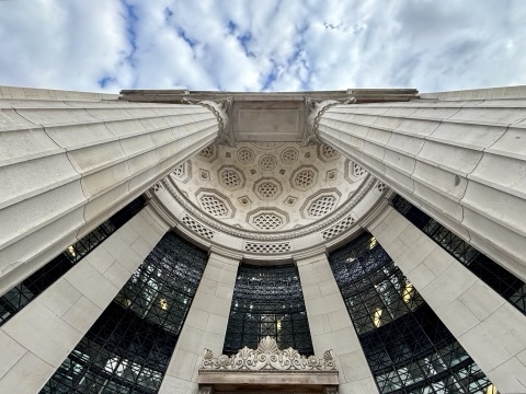 Looking straight up between the fluted stone columns of Bush House's portico to a coffered half-dome, with a glazed screen and a carved shell above the doorway below.