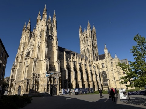 Canterbury Cathedral from the south-west on a clear day: the twin west towers, the long nave and the tall central tower, all in pale stone, with a few visitors on the paved precinct.