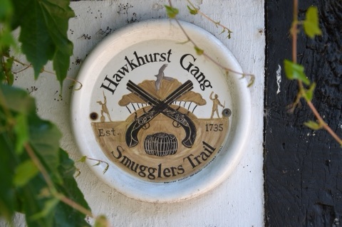 A round painted plaque on a whitewashed timber wall, with crossed flintlock pistols over a barrel, two figures with cutlasses and the words Hawkhurst Gang Smugglers Trail, Est. 1735, half hidden by vine leaves.