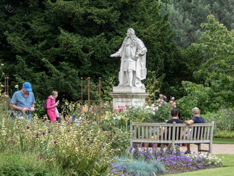 A white marble statue of a man in a long wig and robes on a plinth among flower beds, two people on a wooden bench in front of it and others walking through the planting.
