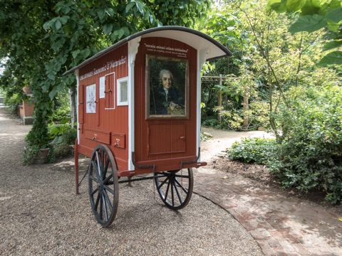 A small red-painted wooden caravan on two spoked wheels, standing on a gravel path under trees, with a framed portrait of Carl Linnaeus on its door and his name on the side.