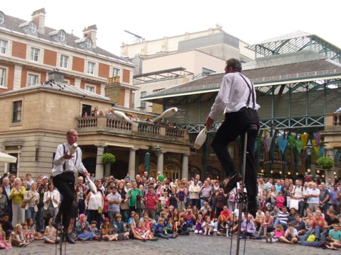 Two jugglers performing on the cobbles of the piazza to a crowd sitting and standing in a ring, one of them balanced on a tall unicycle, with the market building's iron and glass roof and a stucco terrace behind them.