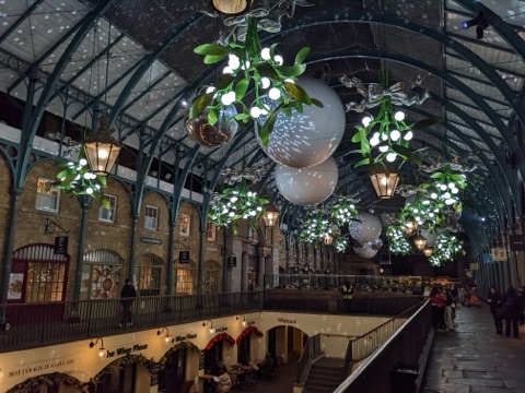 Inside the market hall: a long glazed roof on slender iron columns tied by open arched trusses, brick arcades of shops on both floors below, and large illuminated mistletoe and bauble decorations hung the length of it.