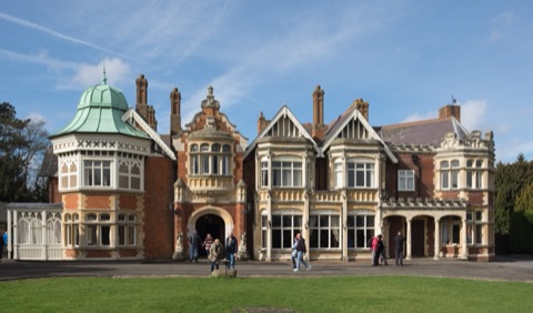 A rambling Victorian mansion of red brick and pale stone with a green copper dome on one corner, bay windows and tall chimneys, visitors crossing the lawn in front.