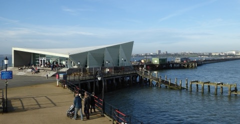 The angular grey pavilion at the end of Southend Pier, people on its terrace, and the pier running away on timber legs across blue water to the town on the shore.