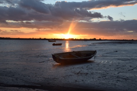 A rowing boat lying on wet mud at low tide, the sun setting behind a low shore with small boats at anchor and the sky breaking orange through dark cloud.