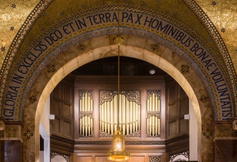 A marble arch inscribed in mosaic letters with Gloria in excelsis Deo et in terra pax hominibus bonae voluntatis, framing the organ's pipes in a carved wooden case, with a brass lantern hanging in front.