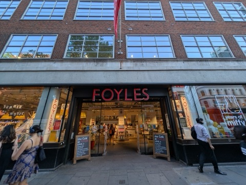 Foyles' red-brick front on Charing Cross Road at dusk: the name in red letters over the open glass doors, lit bookshelves inside, a café board on the pavement and people walking past.