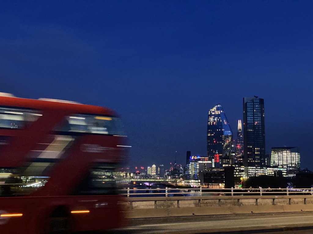 A red double-decker bus blurred with speed crossing Waterloo Bridge at dusk, with the OXO Tower's lit sign and the dark glass curve of One Blackfriars beyond the river under a deep blue sky.
