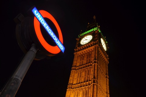 An illuminated London Underground roundel on its pole at night, with the clock tower of the Houses of Parliament lit up behind it