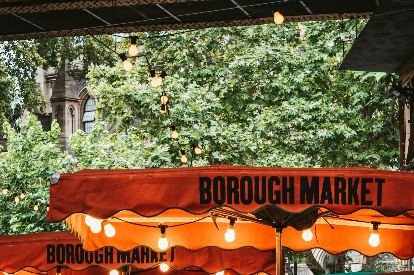 Orange canvas awnings printed with the words Borough Market, strung with lit festoon bulbs, under plane trees with a Gothic stone church behind