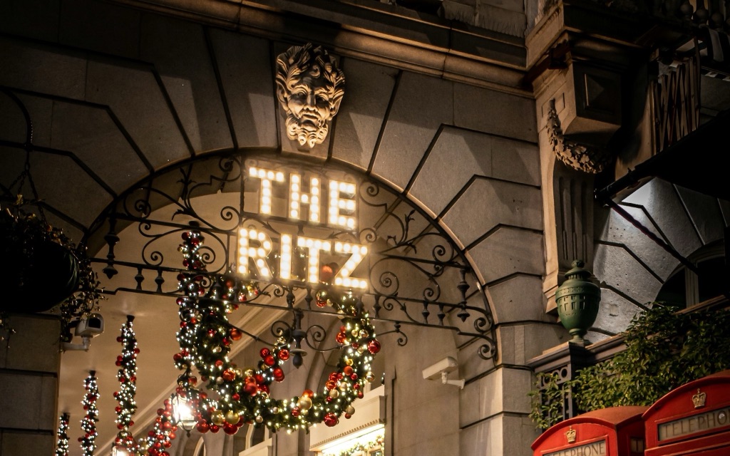 The Ritz's arcade at night: THE RITZ spelled in light bulbs inside an iron arch with a carved mask above it, Christmas garlands of baubles and lights strung along the colonnade, and the crowned tops of two red telephone boxes at the right.
