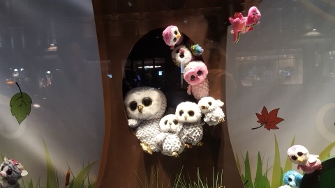 A shop window dressed as a tree trunk with a hollow in it, filled with soft-toy owls and pink and white birds with big glitter eyes, on a bed of paper grass.