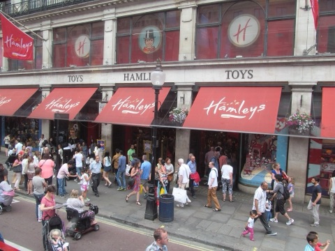 Hamleys' stone front on Regent Street with TOYS carved over the windows, a row of red awnings with the name in white script, red banners above, and a crowd of families on the pavement.