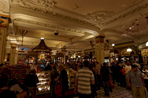 Harrods' food hall: an ornate white plaster ceiling with moulded swags over tiled columns, a pagoda-roofed counter of tea and coffee in the middle, and crowds of shoppers between the counters.