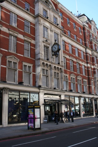 Harvey Nichols' red-brick and stone front on Knightsbridge: tall windows in stone frames, a black and gold clock projecting over the entrance canopy, and shoppers on the pavement by a phone box.