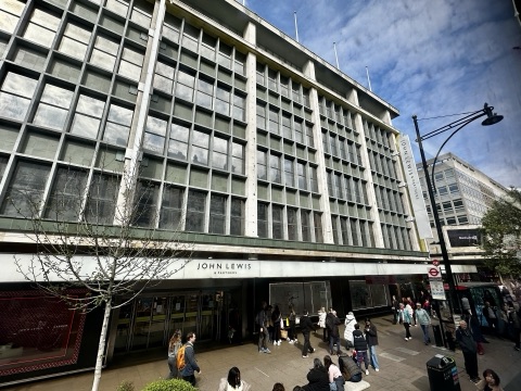 John Lewis's 1960 front on Oxford Street from across the road: a grid of tall windows in concrete frames above the shop's fascia, a young tree in front and shoppers on the pavement.