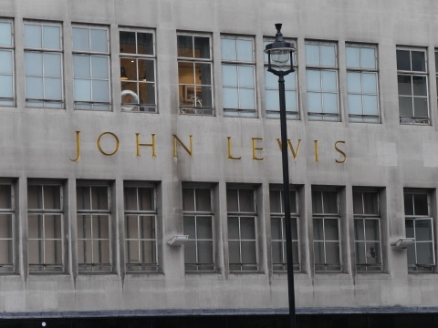 The name John Lewis in gilt letters across a plain Portland stone front between two rows of windows, with a black street lamp in front.