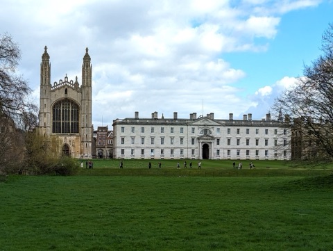 King's College Chapel's pinnacled west end and the long white classical range beside it, seen across a broad lawn from the Backs, with people on the path in front.