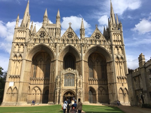 Peterborough Cathedral's west front: three huge pointed arches between spired turrets, a small porch set in the middle one, with visitors on the lawn below.