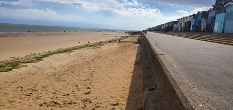 A long sandy beach at low tide beside a concrete promenade, with a row of pastel beach huts running away along the top of the sea wall.