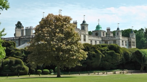The turrets, chimneys and stone parapets of Audley End House rising behind a spreading tree and a bank of clipped yews, under a summer sky.