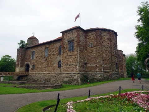 Colchester Castle's great Norman keep of rubble and red Roman brick, with a flag flying, a wooden bridge to its door and flower beds in the park in front.