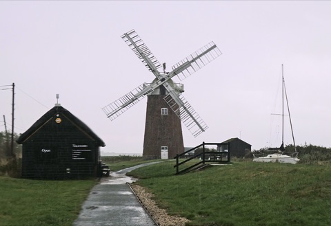 A brick windpump with white sails beside a marsh, a black tarred boathouse marked Open, and a sailing boat moored in the reeds under a grey sky.