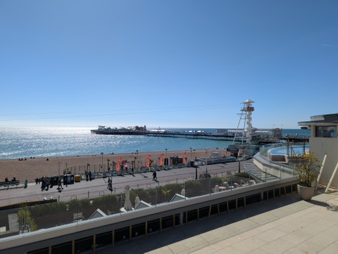 Brighton's pier stretching out over a glittering sea from a shingle beach, with a viewing tower and beach bars along the front in bright spring sun.