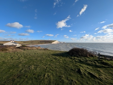 A row of coastguard cottages on a green clifftop above a bay, with the white cliffs of the Seven Sisters beyond under a blue sky and scattered cloud.