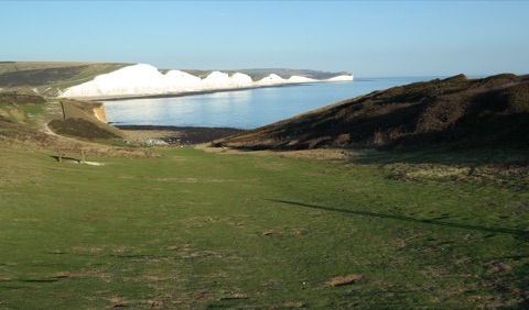 The white chalk cliffs of the Seven Sisters catching low sun across a calm bay, seen from a grassy valley leading down to the shingle.
