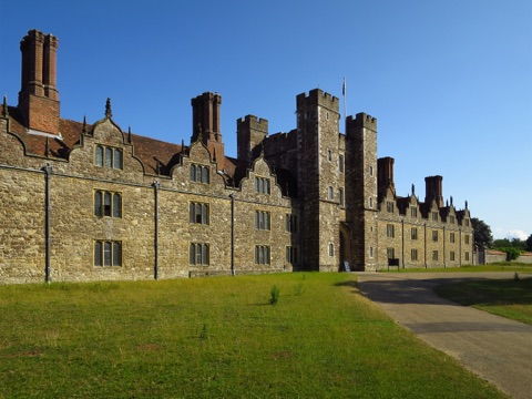 Knole's long stone front of Dutch gables, tall brick chimneys and a battlemented gatehouse tower, on a lawn in evening sun.