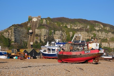 Two fishing boats hauled up on a shingle beach, one red-hulled, in front of black net huts and the funicular railway climbing the sandstone cliff behind.