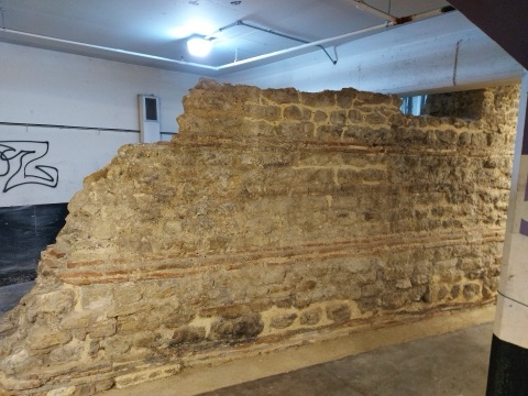 A section of Roman wall in Kentish ragstone with bands of red tile running through it, standing against the painted blockwork and strip lights of an underground car park.