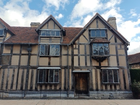 A timber-framed house with leaded windows, two gables and a low wooden door, behind iron railings on a pavement, under a blue sky with white clouds.
