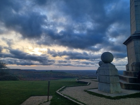 The stone base of a monument on a hilltop at dusk, with a gravel path and a wide view over the Vale of Aylesbury under dark clouds breaking to the west.