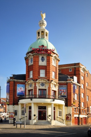 The theatre on a street corner: a red brick and cream stone front under a green copper dome with NEW WIMBLEDON THEATRE across it in red neon, a gilded winged figure on a white globe at the very top, 1910 carved below the dome, a curved colonnaded entrance with red doors at street level and show posters along both flanks.