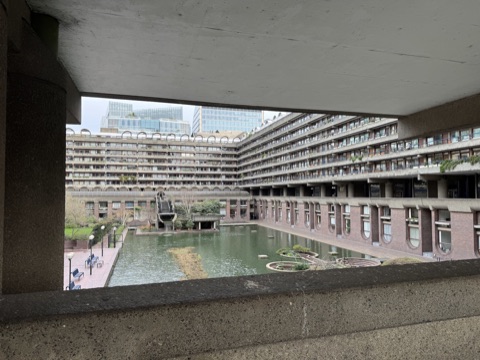 The Barbican's lake seen from under a concrete walkway: a long block of flats with rows of balconies curving round the green water, a waterfall spilling from a raised platform, benches on the brick terrace and glass towers beyond.