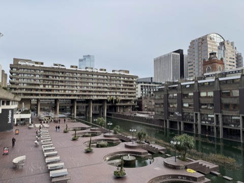 The lakeside terrace in winter: rows of white benches on brick paving, round brick planters with fountains at the water's edge, a long terrace block on stilts across the lake, and a church tower among the towers of the City behind.