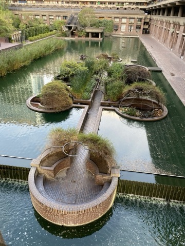 Looking down on the lake: a round brick platform at the edge of a weir in front, a brick walkway leading out to planted islands of reeds and shrubs under iron domes, and the terrace blocks with their curved balconies around the water.