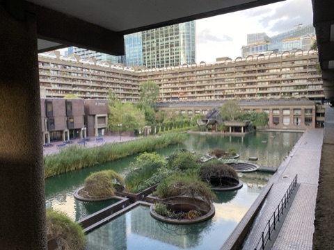 The lake from under the high walk: planted islands and reeds in green water, a brick terrace with benches, a long block of flats with rows of balconies enclosing the far side, and glass towers rising behind it.