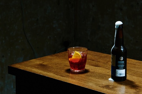 A negroni in a tumbler with an orange slice, beside a beer bottle, on a wooden table against bare concrete