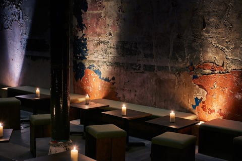 Candlelit tables and low green stools along a basement wall of bare plaster, peeling to patches of orange and blue
