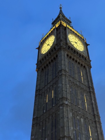 The top of the Elizabeth Tower against a deep blue evening sky, two of its clock faces lit gold, with the gilded ironwork and spire above them.