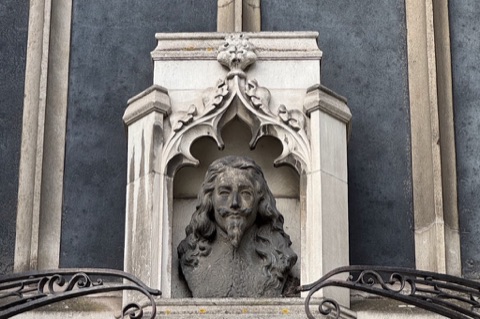 A weathered lead bust of Charles I, long-haired and bearded, in a carved Gothic stone niche on a church wall, with dark ironwork below it