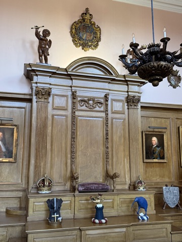 A raised wooden throne with a velvet cushion under a carved canopy, a gilt crown on either side, painted heraldic crests on the bench below, a cherub with a baton and the royal arms above, portraits on the panelled walls and a carved chandelier.