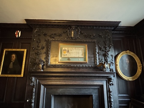 Dark carved panelling over a doorway: swags of flowers and leaves around a framed document under glass, a gilt eagle and a small shield on the shelf, a portrait in a gold frame to the left and an oval one to the right.
